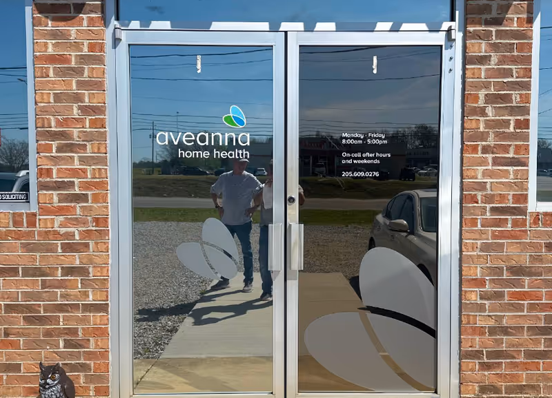 Glass double doors with Aveanna Home Health logo, brick exterior, and reflection of a person outside.