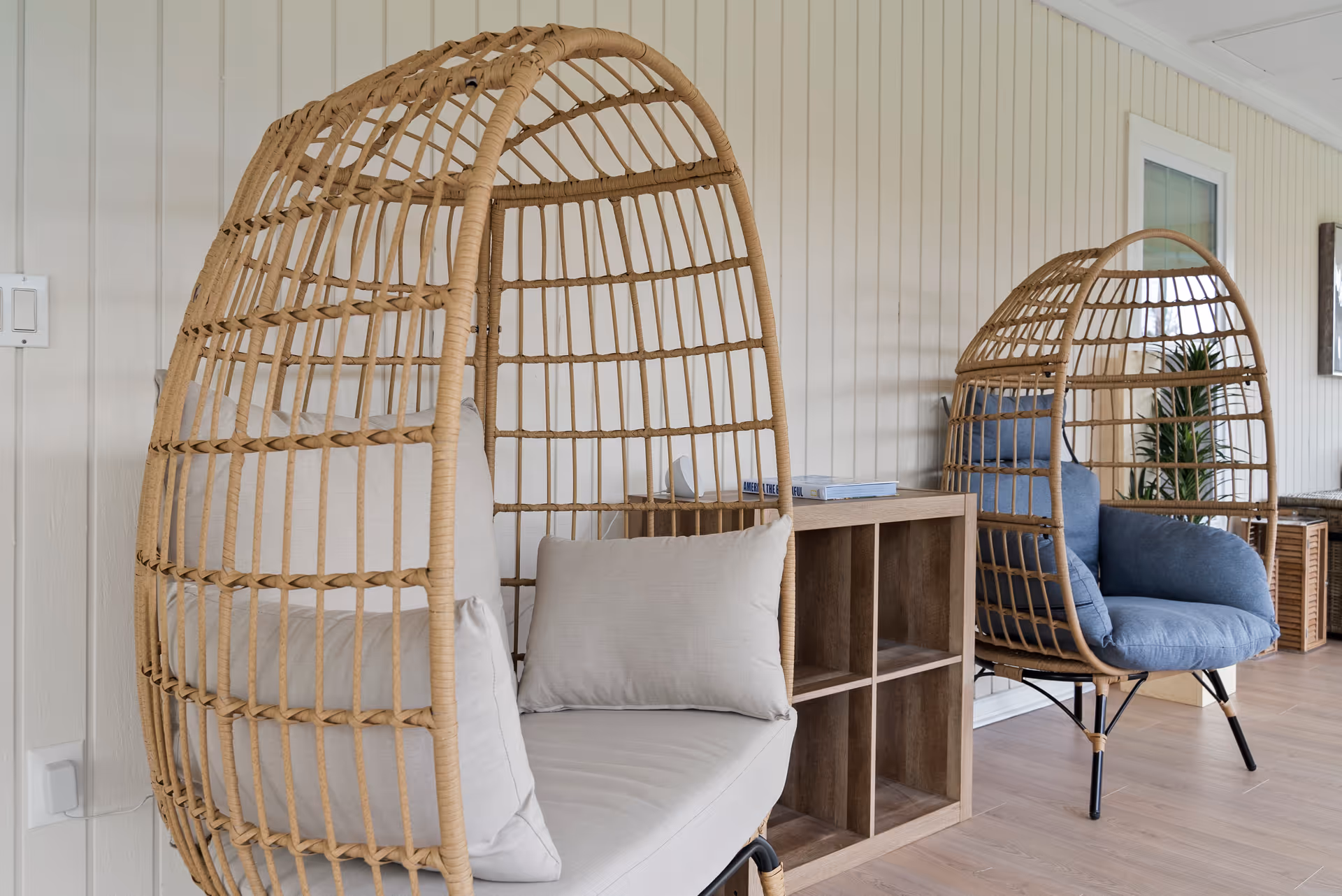 Two wicker egg chairs with cushions sit beside a cubby shelf in a bright interior.