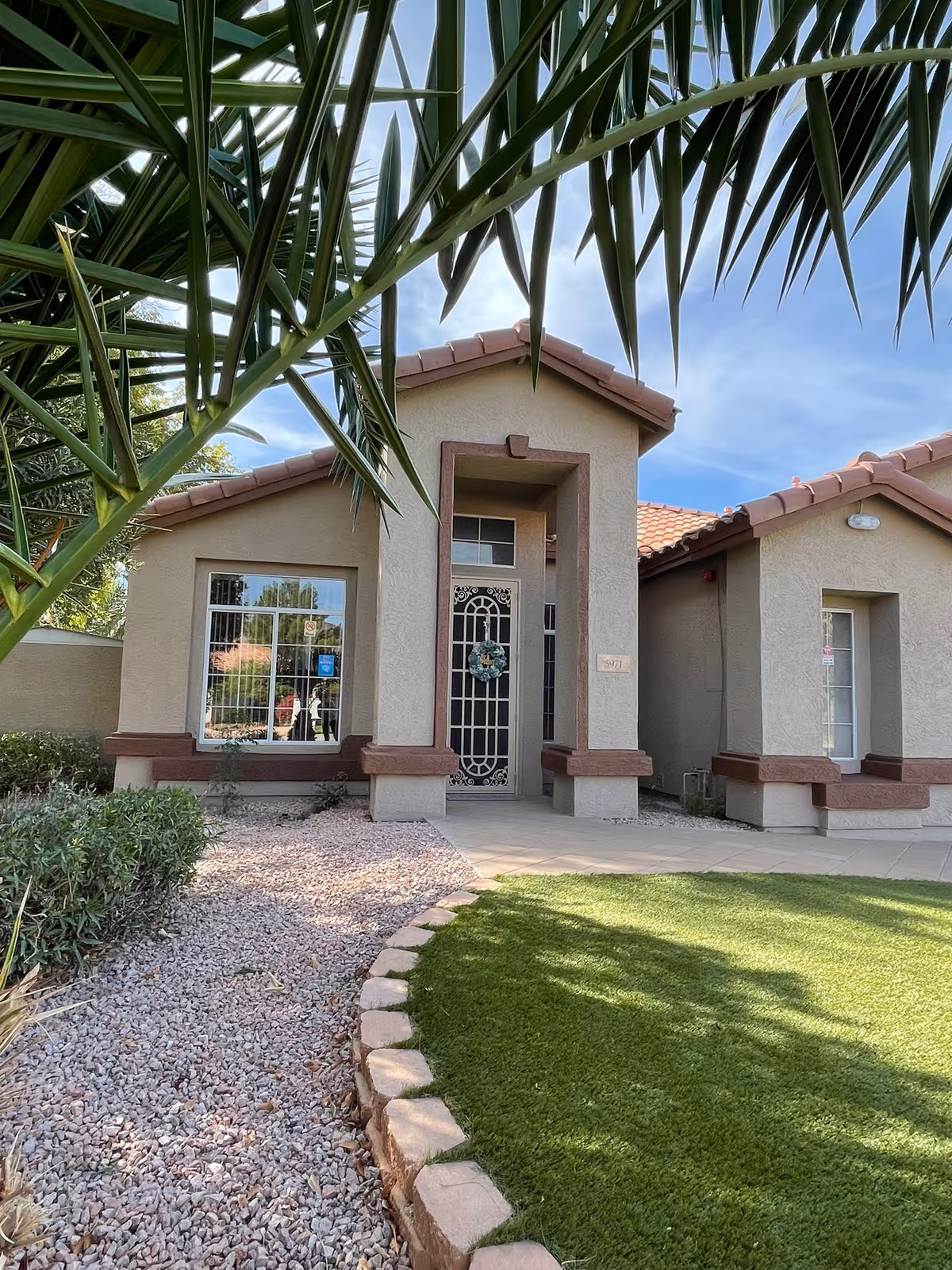 Front exterior of a single-story stucco building with a decorative metal entry door, wreath, tiled roof, and landscaped yard with rocks and artificial turf.
