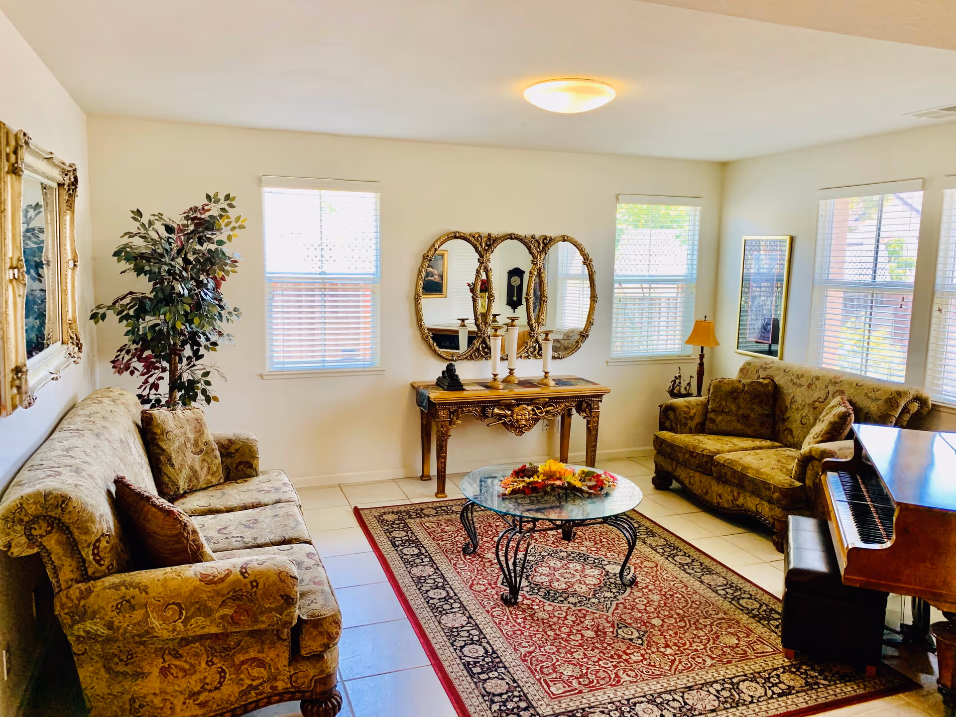 A bright living room with two patterned sofas facing each other, a glass coffee table with a floral centerpiece on a red and beige patterned rug, a decorative console table with three ornate mirrors above it, a floor lamp, a potted plant, and a piano with a bench near the windows.