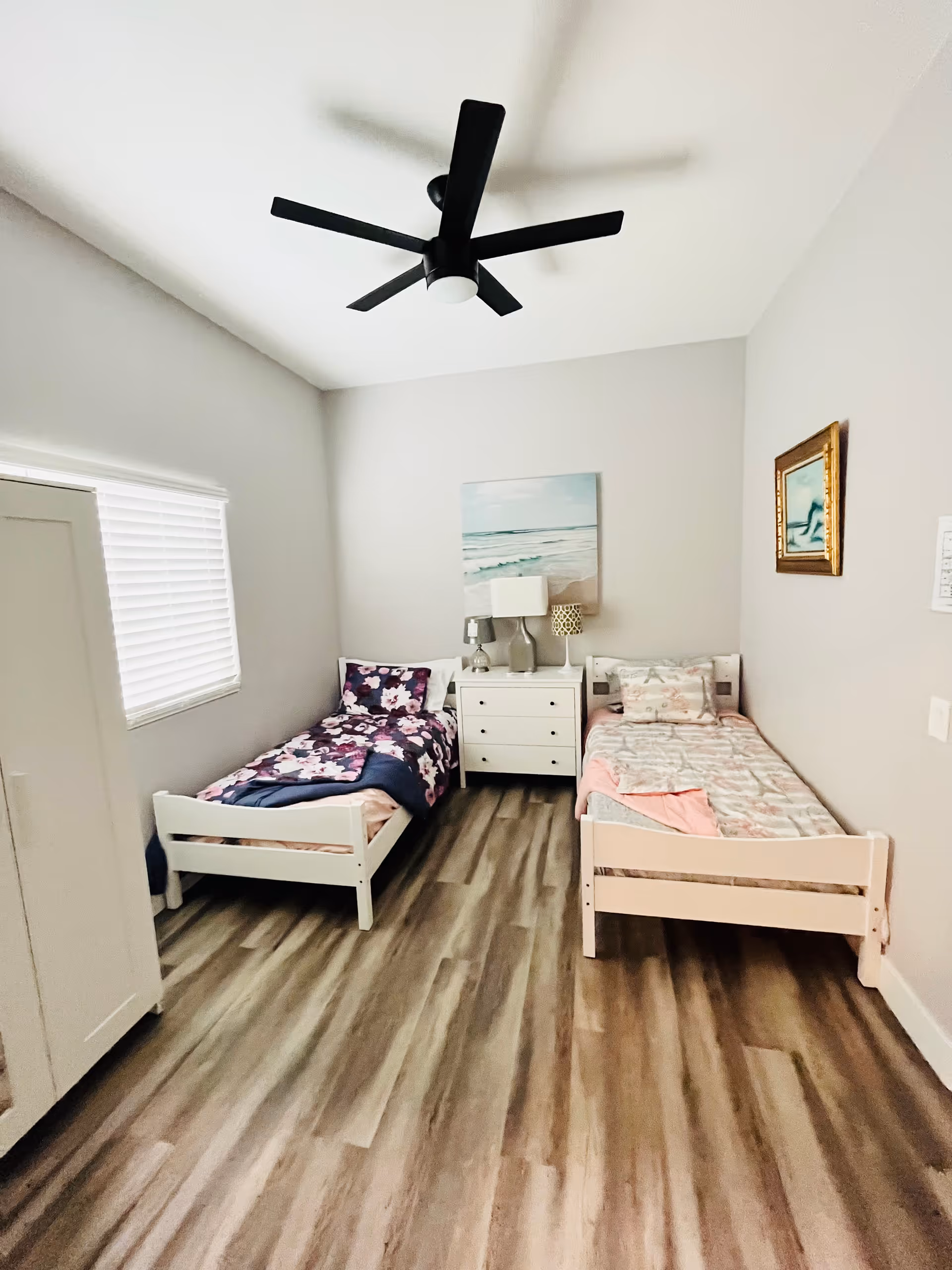 A small bedroom with two single beds placed parallel to each other with a white nightstand in between. The bed on the left has a floral purple and pink bedspread, while the bed on the right has a light-colored patterned bedspread. Above the nightstand is a painting of a beach scene. The room has light gray walls, a window with white blinds on the left wall, a ceiling fan with black blades, and wood-look flooring.