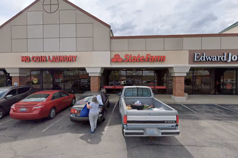 Person unloads items from a blue bag into a car in a parking lot by storefronts.