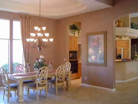 A dining area with a long table, chandelier, and open kitchen in a senior living facility.