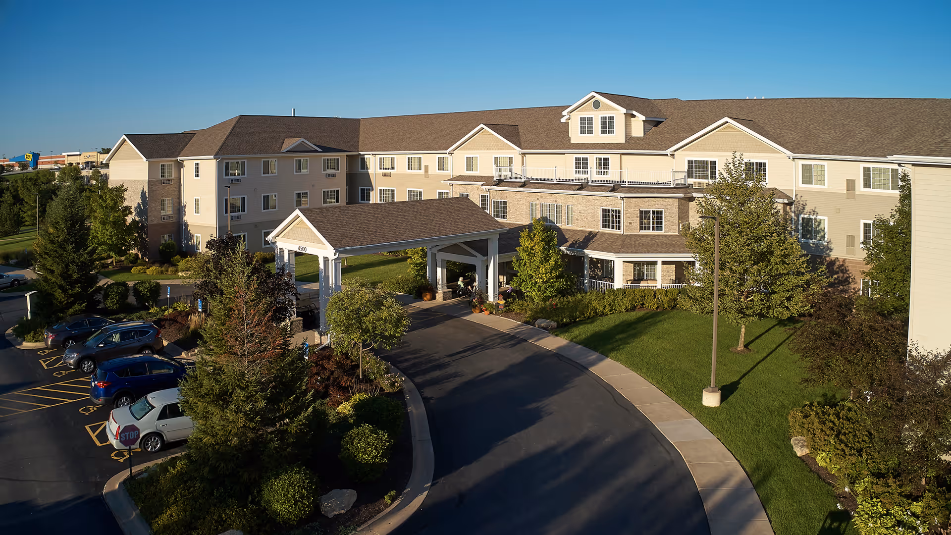 Three-story senior living building with a covered entrance, curved driveway, parked cars, and landscaped grounds under a clear blue sky.
