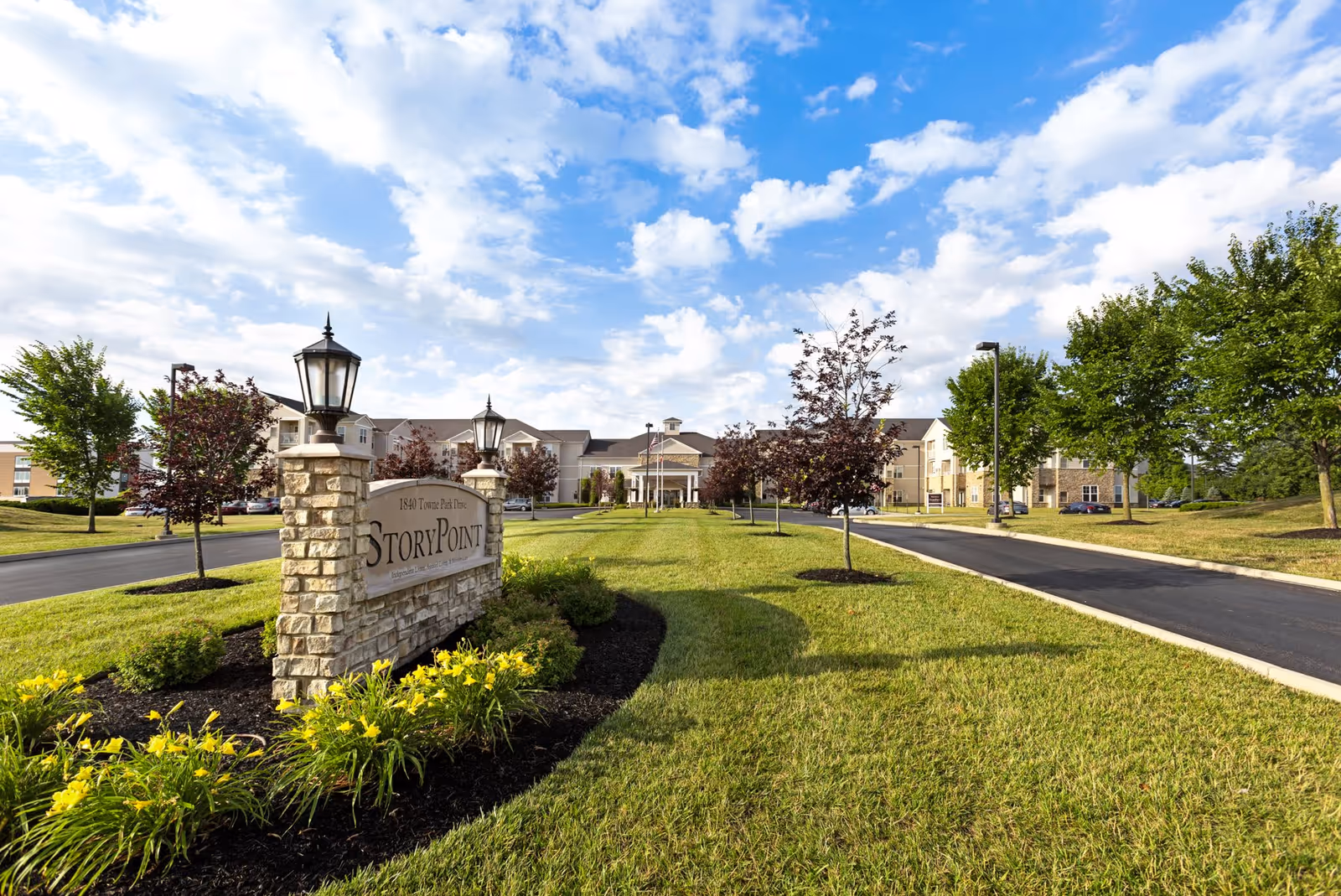 Wide exterior view of StoryPoint Troy senior living facility with a stone sign in the foreground surrounded by yellow flowers and a driveway leading up to the building under a partly cloudy blue sky.
