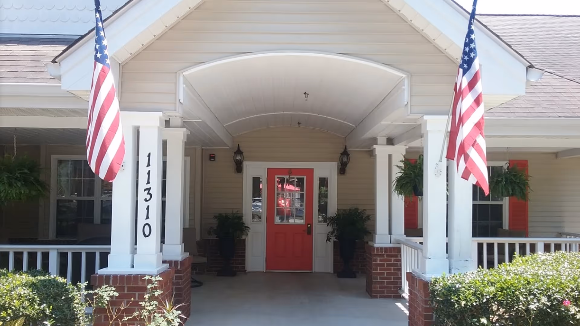 Entrance of a building with a covered porch featuring white columns and brick bases, two American flags hanging on either side, a red door in the center, and green plants placed near the entrance.