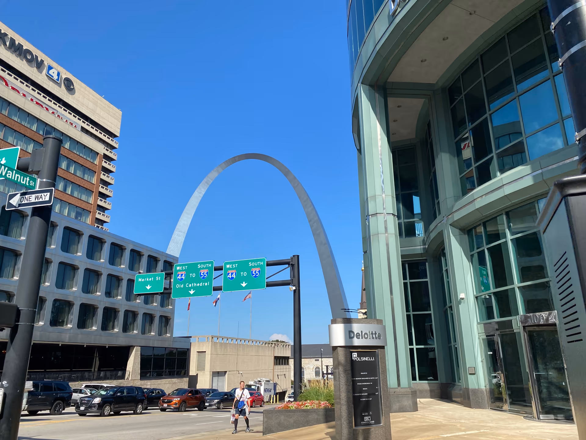 Urban street scene with modern glass building and Gateway Arch in the background.