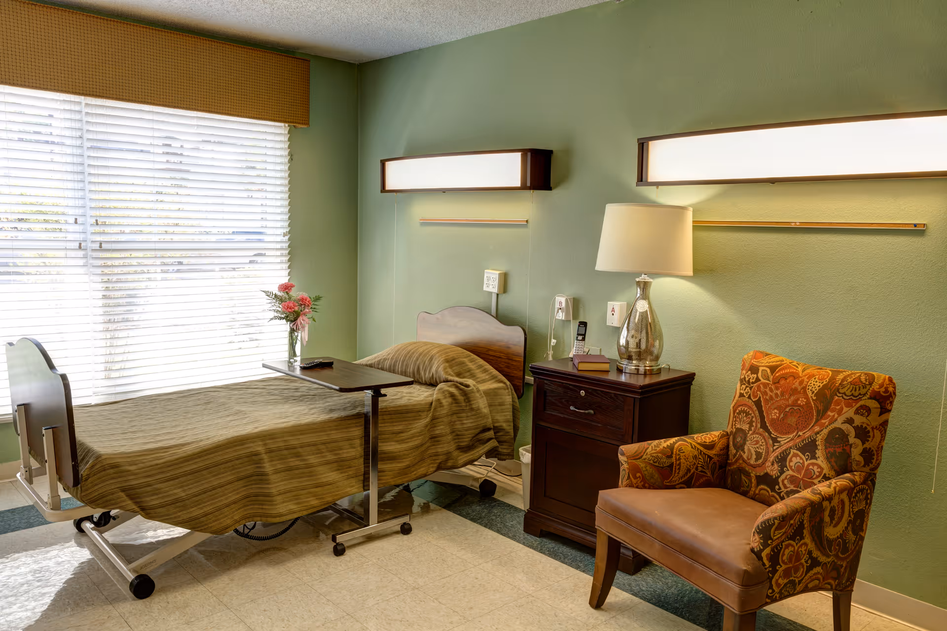 A hospital-style bedroom with a single bed covered in a brown striped blanket, a small overbed table with a vase of flowers, a wooden nightstand with a lamp and books, and a patterned armchair. The room has green walls and a large window with blinds letting in natural light.