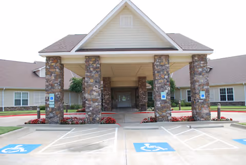 Entrance of a senior living facility with a covered driveway supported by stone pillars, handicap parking spaces in front, and a beige building with a sloped roof in the background.