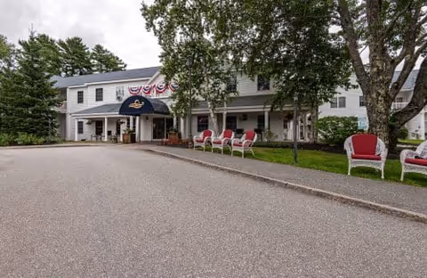 Exterior view of a two-story white building with a covered entrance decorated with patriotic bunting. Several red and white chairs are arranged along the sidewalk under large trees, with a paved driveway in front.