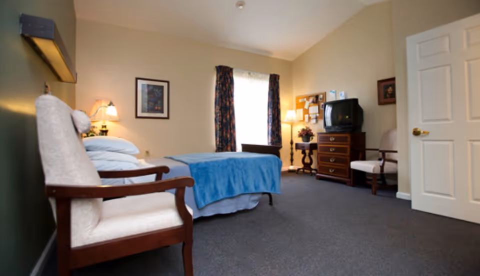 A cozy bedroom with a single bed covered with a blue blanket, two cushioned wooden armchairs, a wooden dresser with an old-style TV on top, a small side table with a lamp and flowers, a bulletin board on the wall, and a window with floral curtains letting in natural light.