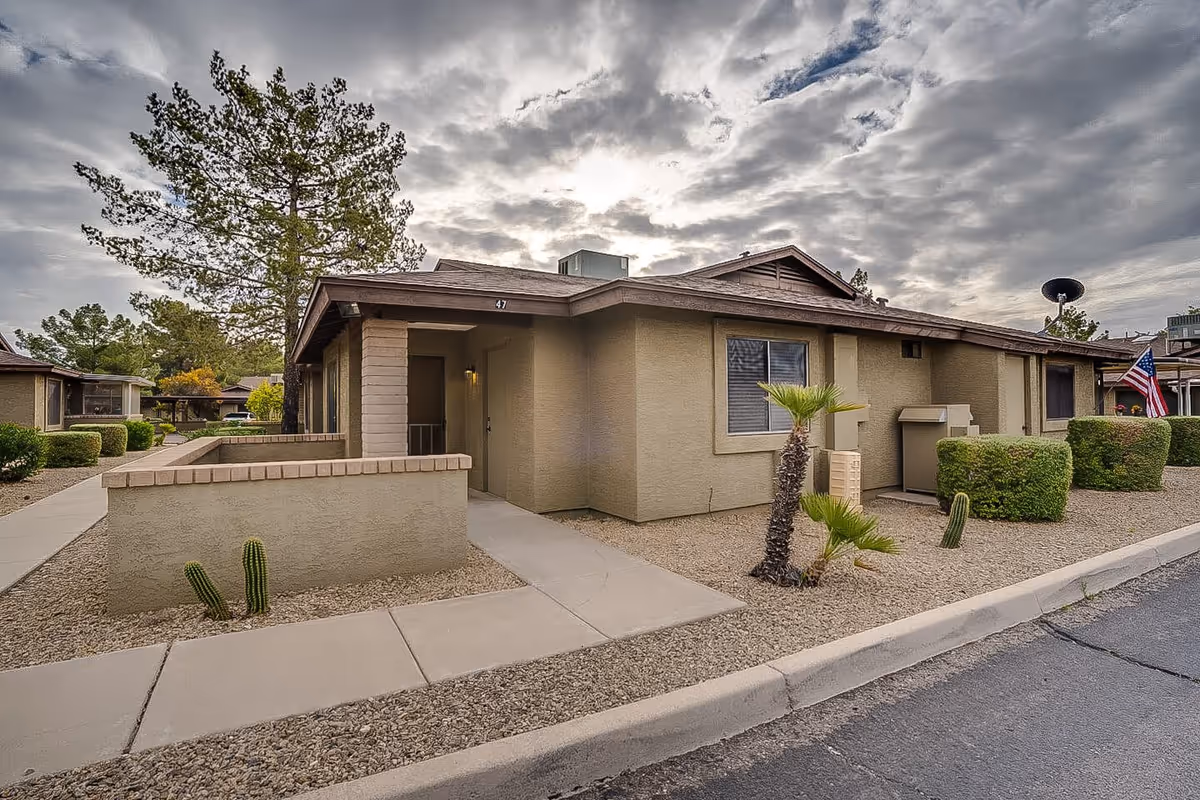 Exterior view of a single-story residential building with beige walls and a brown roof under a cloudy sky. The building has a small enclosed patio area with a low wall, a concrete walkway, desert landscaping with small cacti and bushes, and an American flag visible in the background.