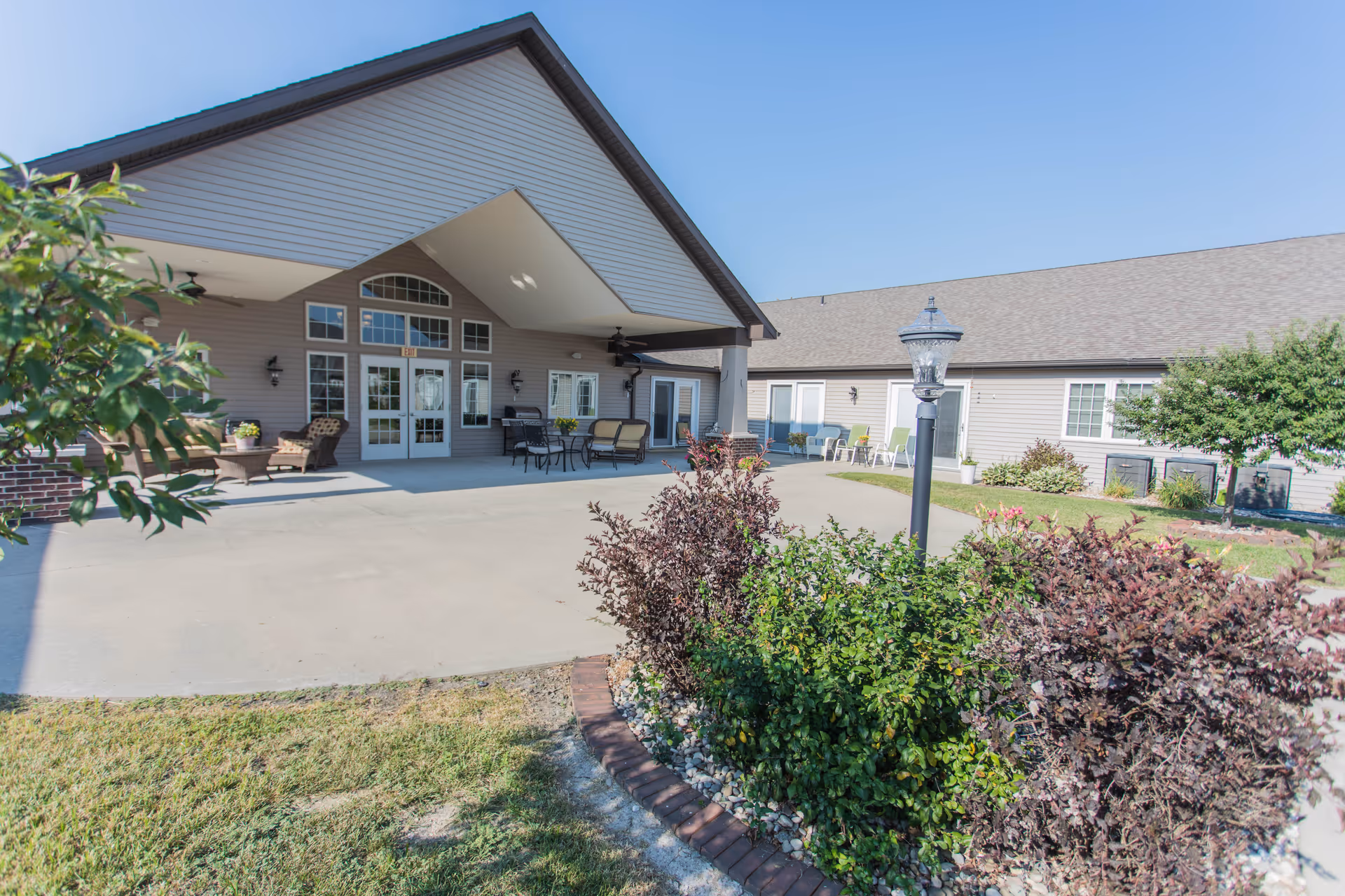 Exterior view of a senior living facility with a covered patio area featuring outdoor seating and tables. The building has beige siding, multiple windows, and a peaked roof. There are landscaped bushes and a lamp post in the foreground under a clear blue sky.