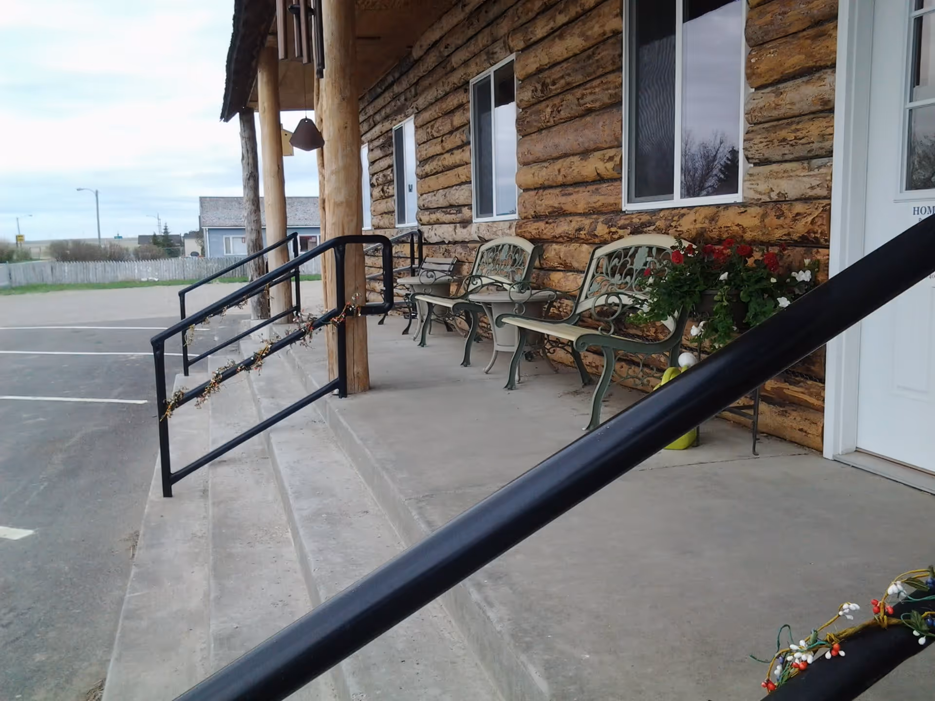 Front porch of a log building with metal benches, potted flowers, and handrails along concrete steps.