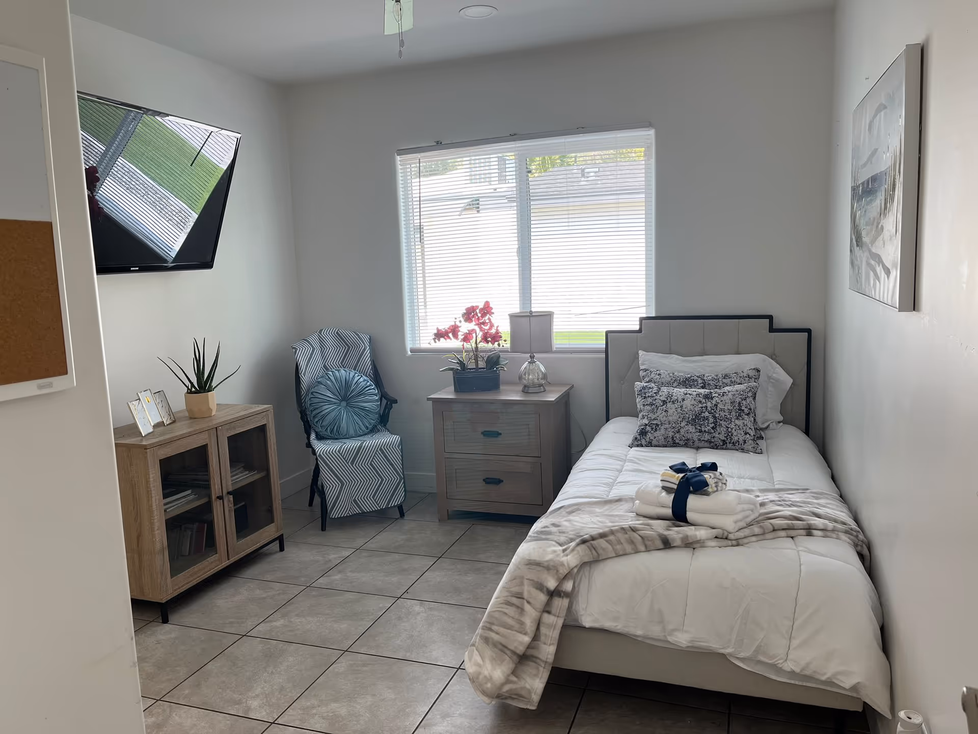 A compact bedroom in a senior living facility featuring a single bed, nightstand, chair, and window.