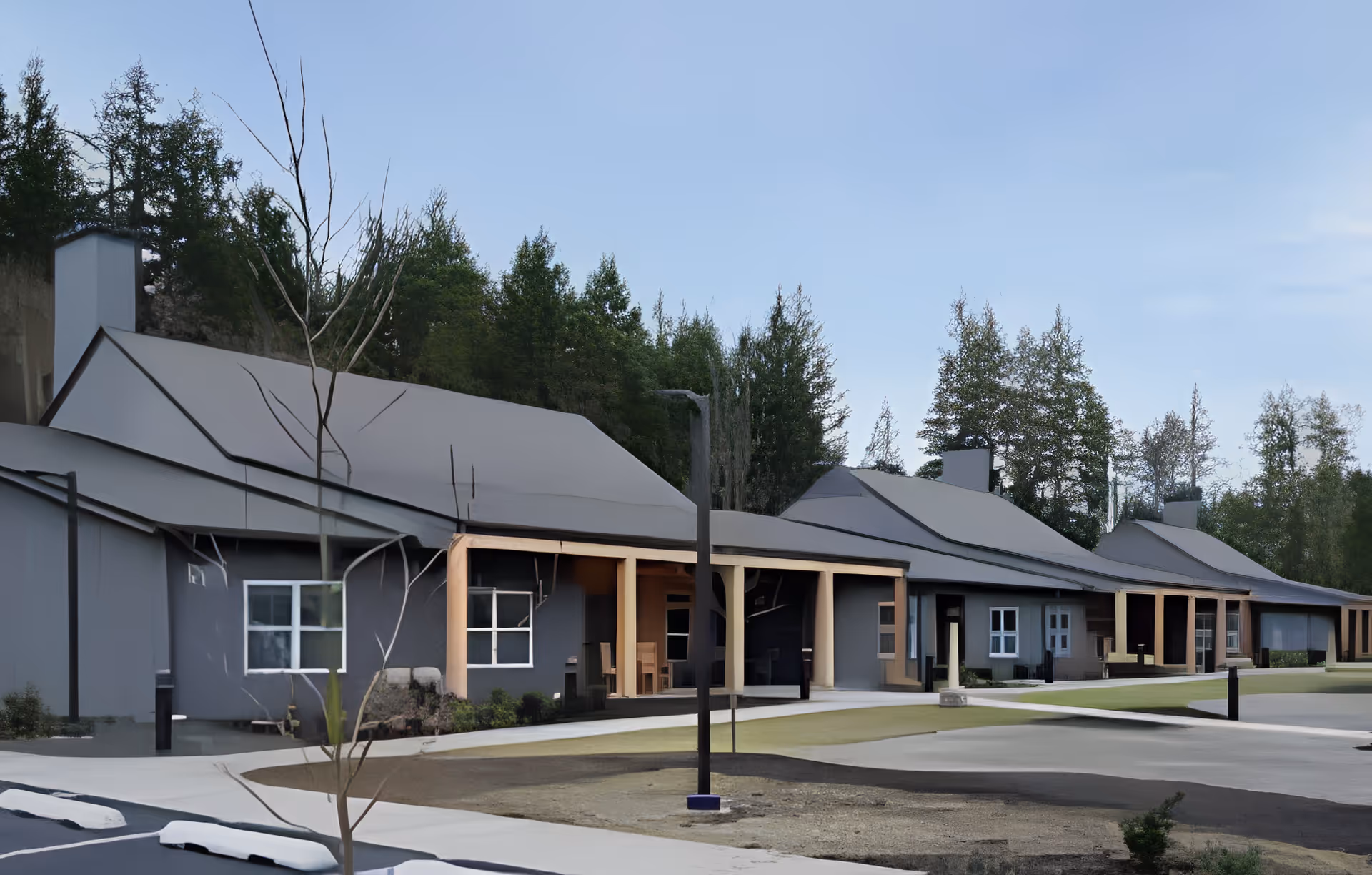 Exterior view of a single-story senior living facility building with multiple units, each having a covered porch area. The building is surrounded by trees and has a paved walkway and parking area in front.
