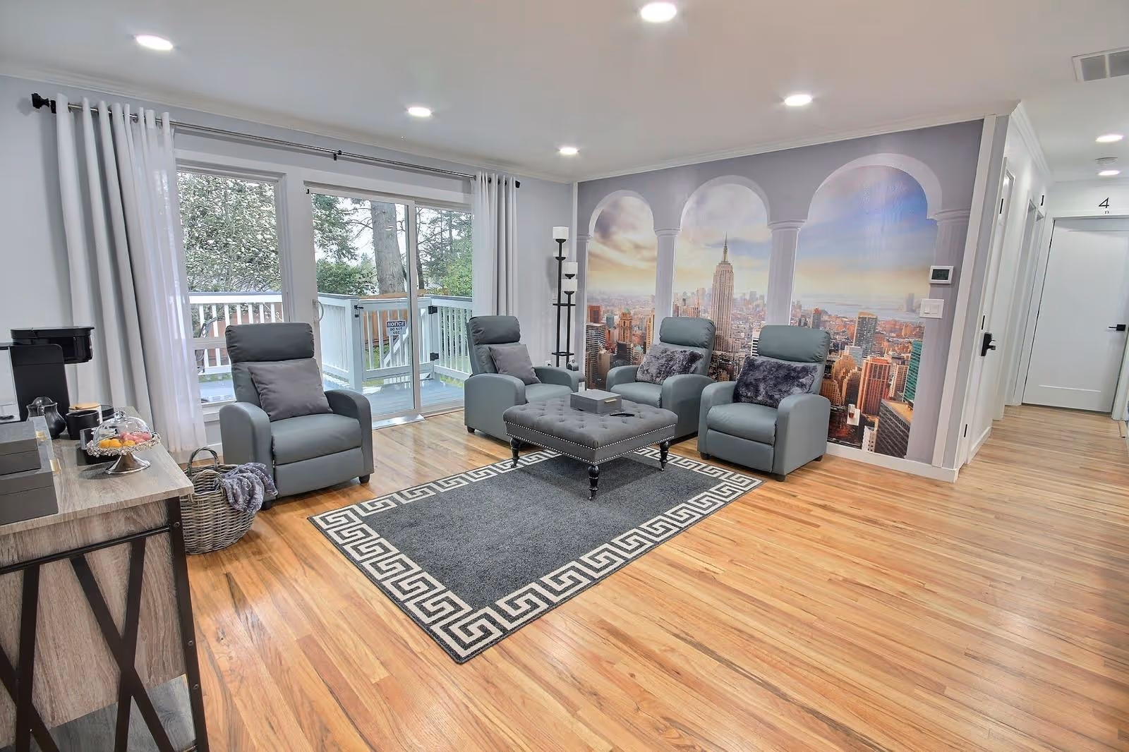 Bright communal living room with four recliners around a central ottoman, overlooking a city mural.