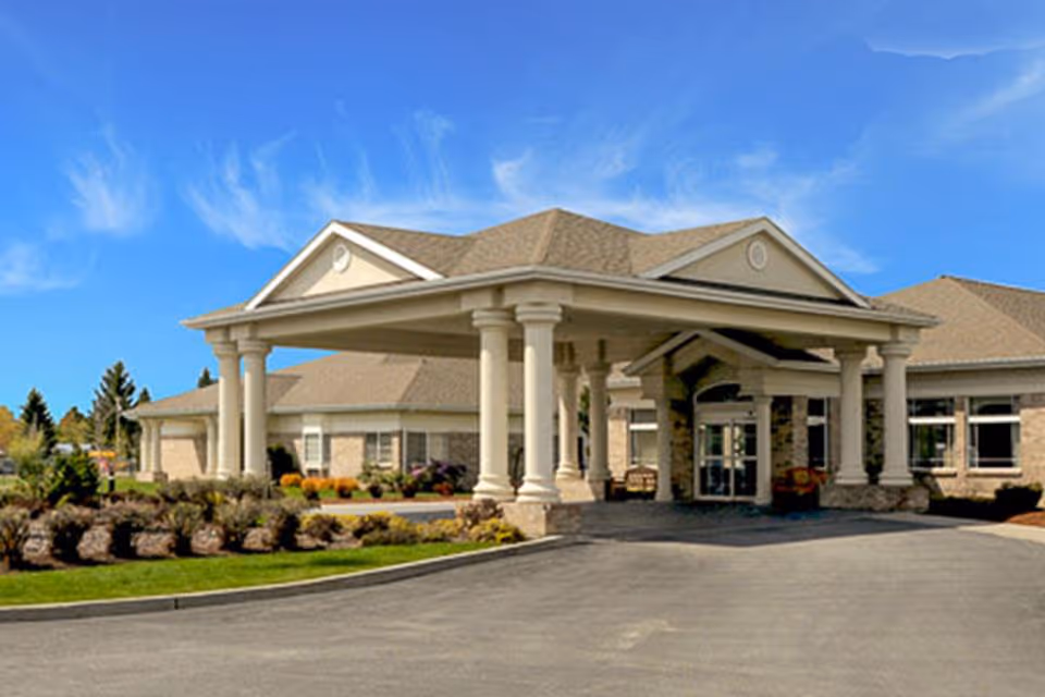 Front exterior view of a single-story building with a large covered entrance supported by white columns, surrounded by landscaped bushes and a clear blue sky.