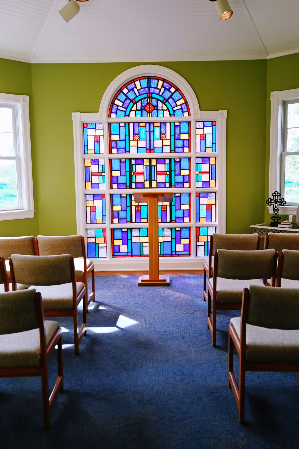 Interior of a small chapel or meditation room with green walls, a blue carpet, rows of wooden chairs with beige cushions, a wooden lectern, and a large colorful stained glass window at the front. Two windows flank the stained glass, and a small table with a cross and books is visible on the right side.