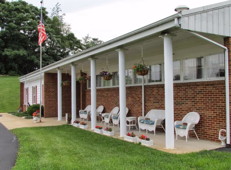 Brick senior living facility front porch with white columns, hanging flower baskets, wicker chairs, and an American flag.
