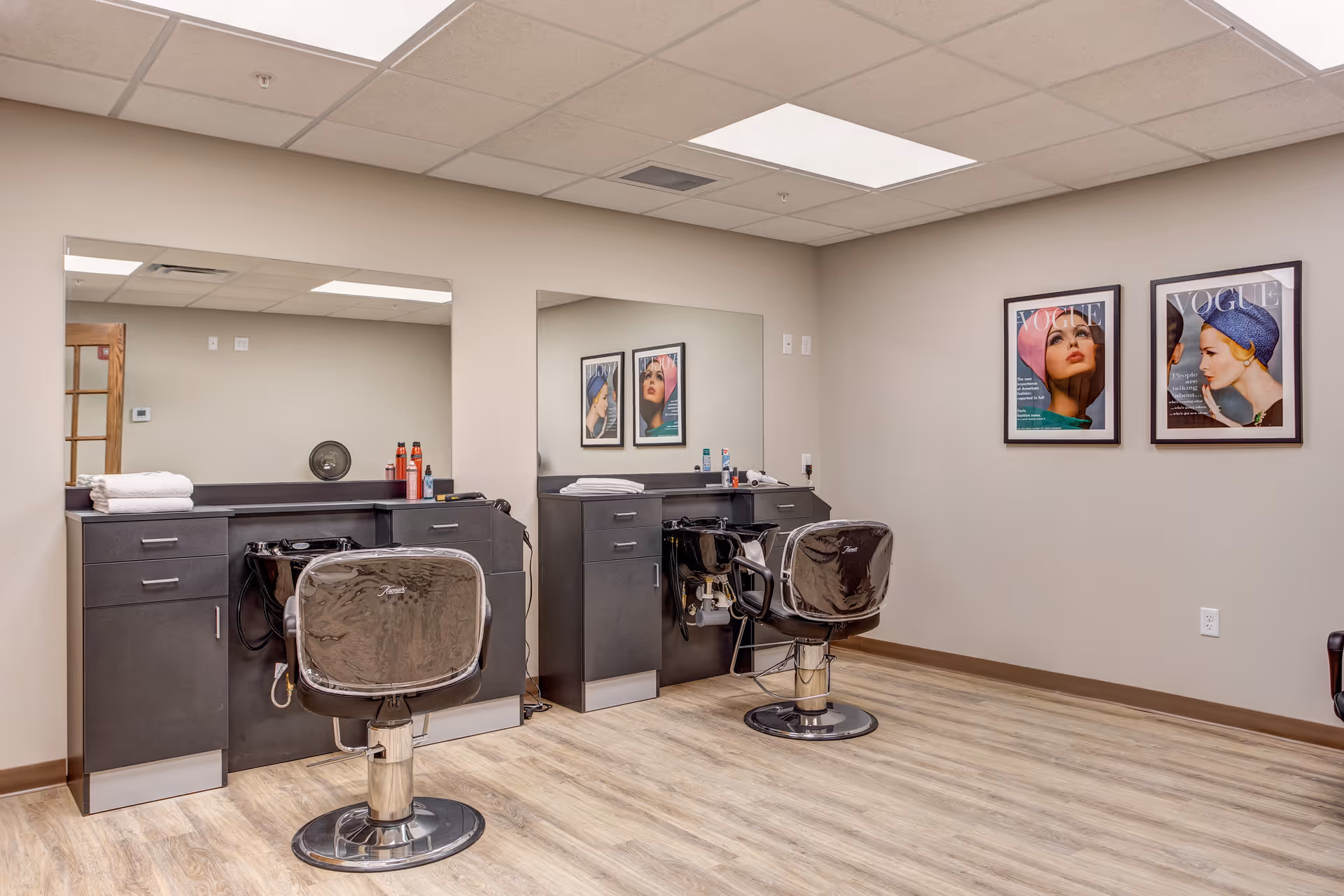 Interior hair salon in retirement community with two styling chairs, mirrors, and framed Vogue posters.