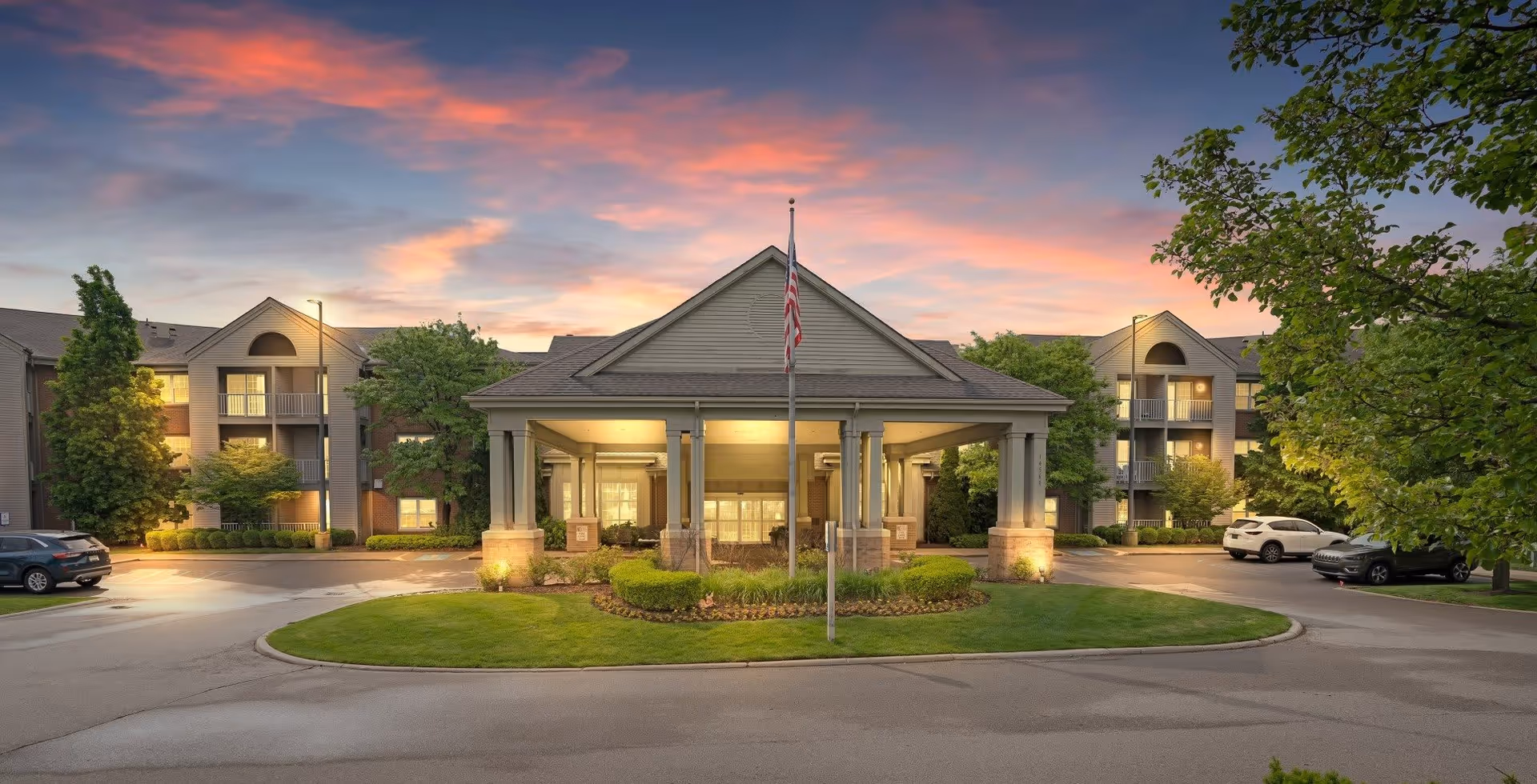 Exterior view of a senior living facility named Shorehaven at sunset, featuring a central entrance with a covered driveway, an American flag on a flagpole, surrounding greenery, and parked cars on either side.