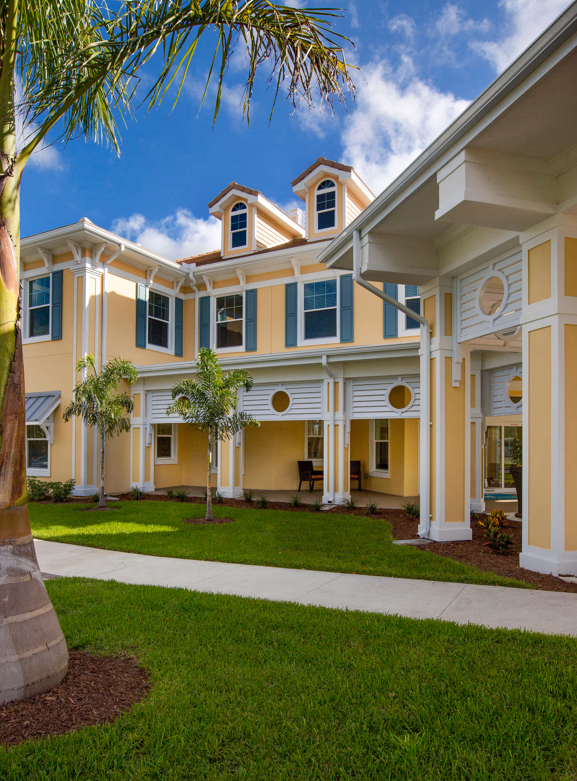 Exterior view of HarborChase of Sarasota, a light yellow senior living building with palm trees.