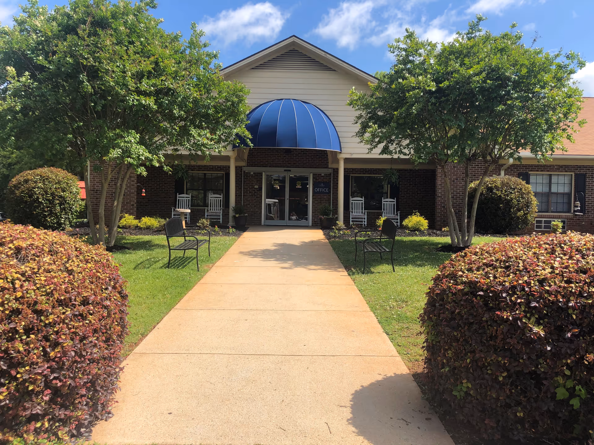 Front exterior view of a single-story brick building with a blue awning over the entrance. There is a concrete walkway leading to the entrance, flanked by green grass, bushes, trees, and benches. The entrance has glass double doors with a sign that reads 'Office'.