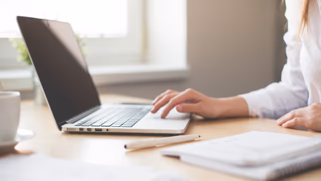 Hands typing on a laptop at a desk with notebook and mug indoors.
