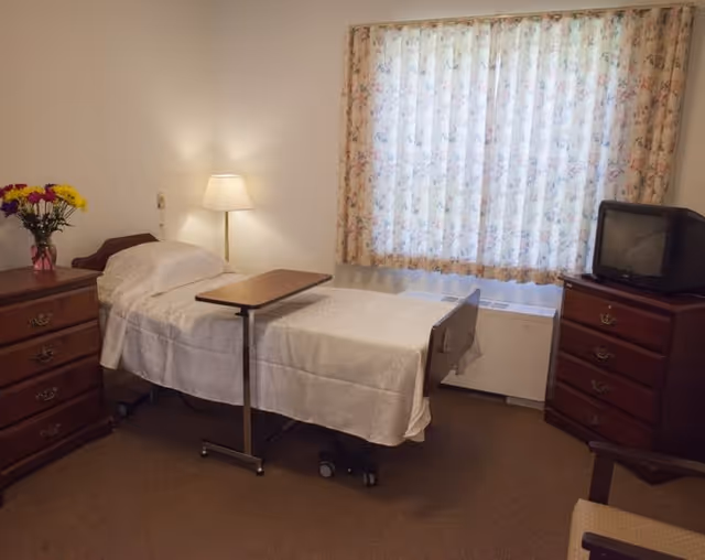A simple senior living bedroom with a single adjustable bed, overbed table, two wooden dressers, a small TV, lamp, and a window with floral curtains.