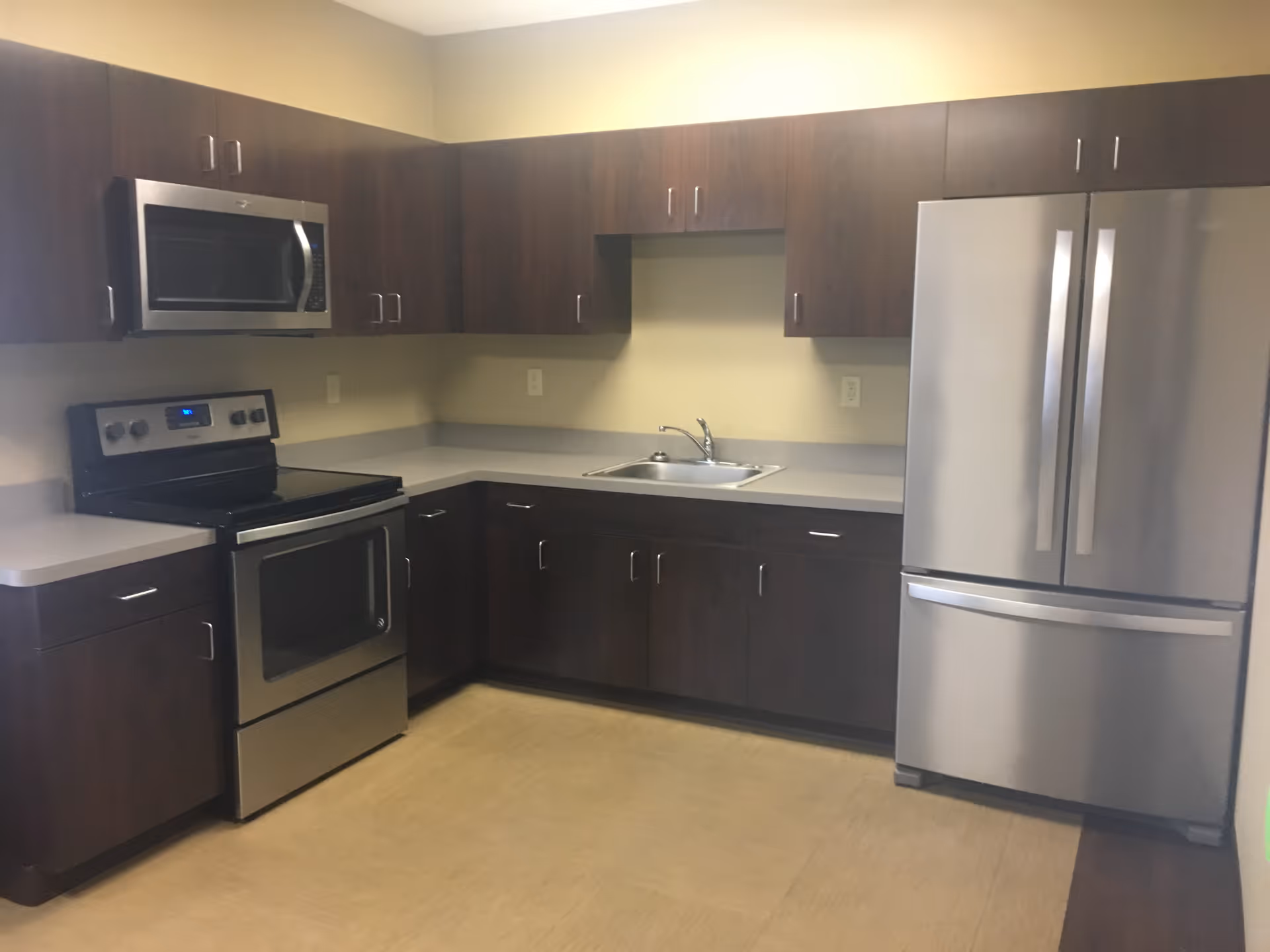 Modern kitchen with stainless steel appliances including a refrigerator, oven, and microwave. The kitchen has dark wood cabinets and light-colored countertops with a sink in the corner.