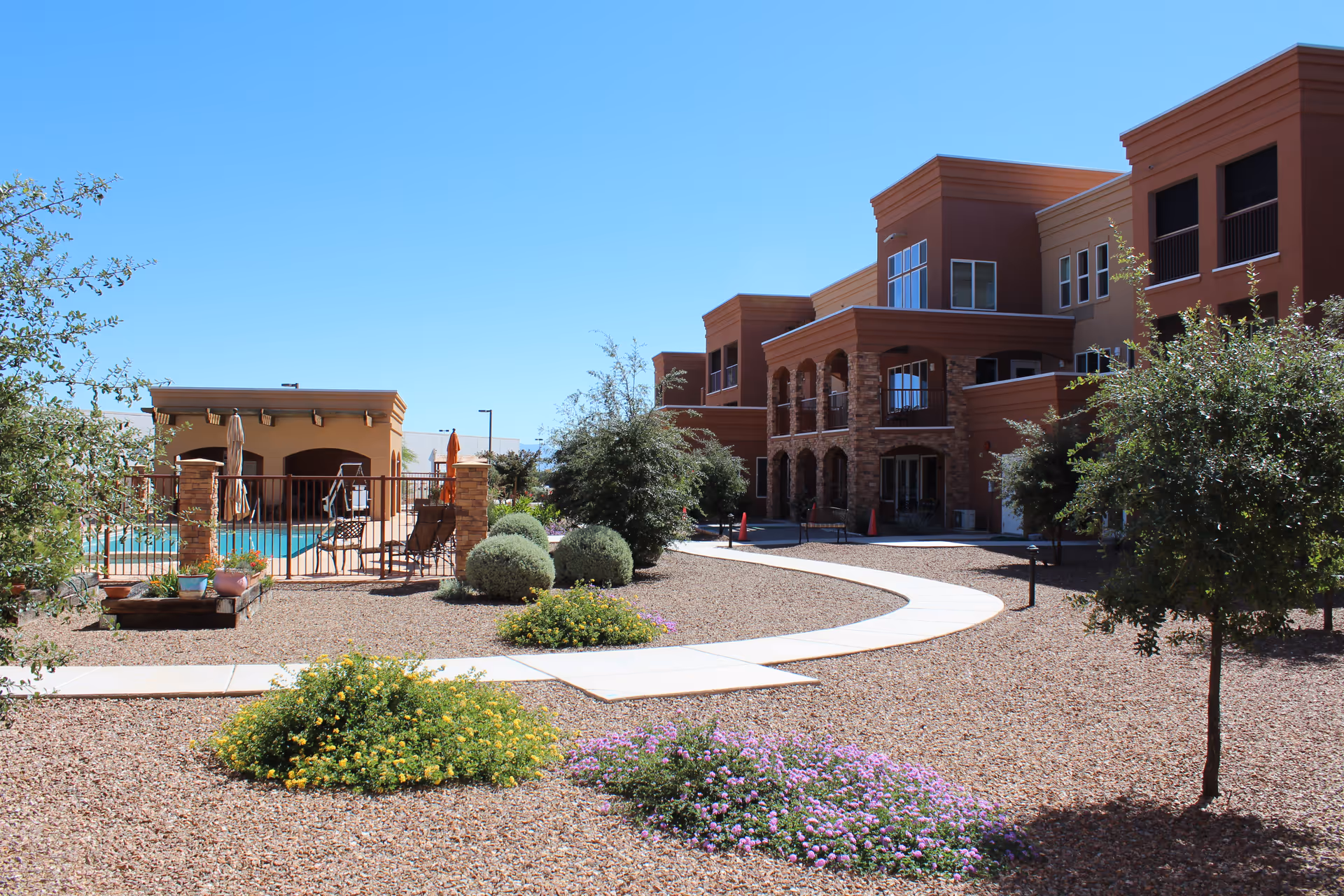 Outdoor view of a senior living facility with a clear blue sky, landscaped garden with bushes and flowers, a curved concrete pathway, and a multi-story building with balconies and stone accents. There is a fenced pool area with lounge chairs and umbrellas on the left side.