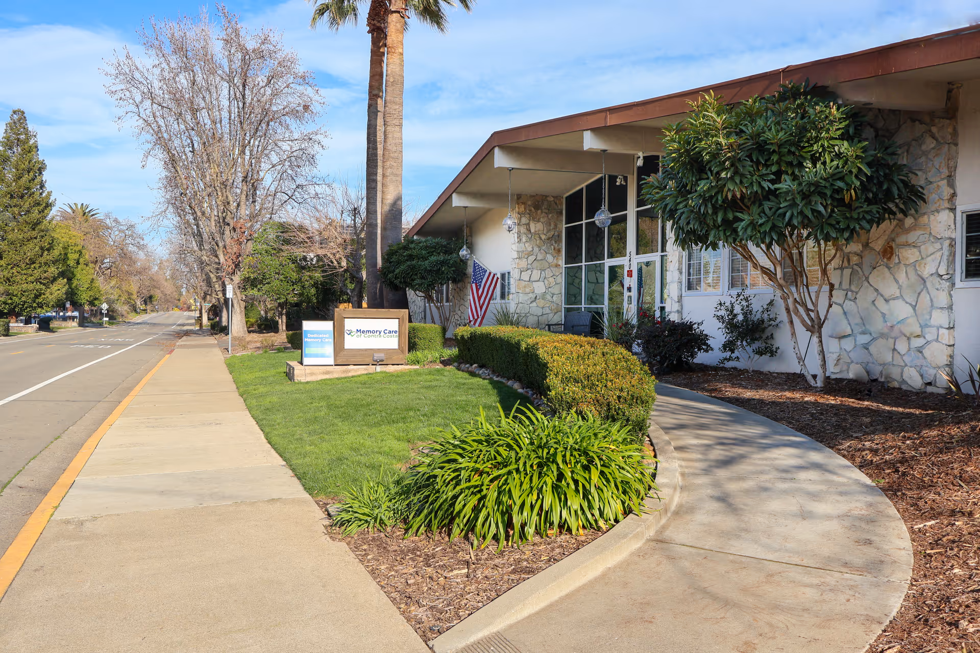 Exterior view of Memory Care of Contra Costa facility showing a sidewalk, green lawn, bushes, trees, and a stone-faced building with large windows and an American flag near the entrance.