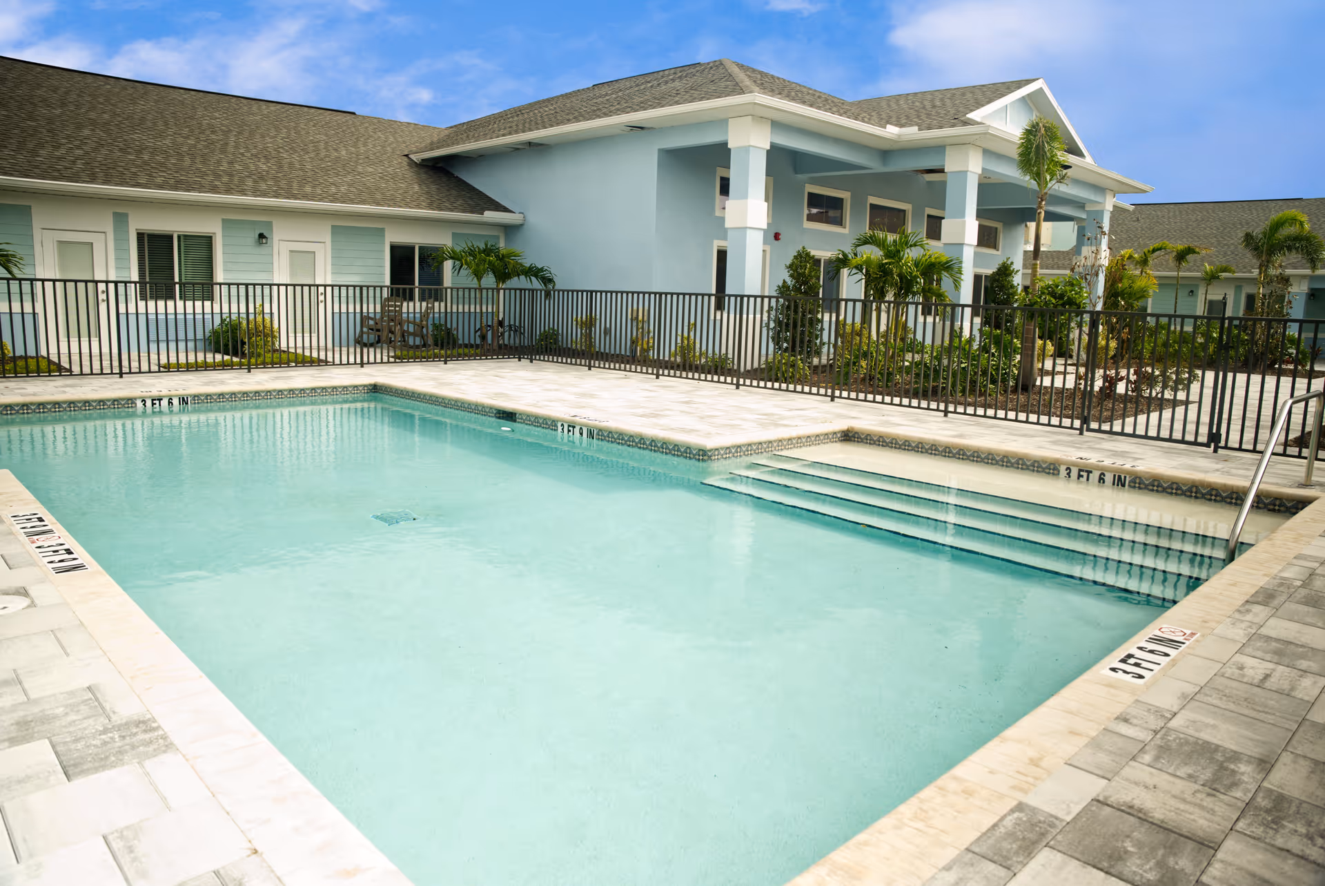 Outdoor swimming pool with clear blue water surrounded by a tiled deck and black metal fence. In the background, there is a light blue building with white trim, palm trees, and some landscaping under a partly cloudy sky.