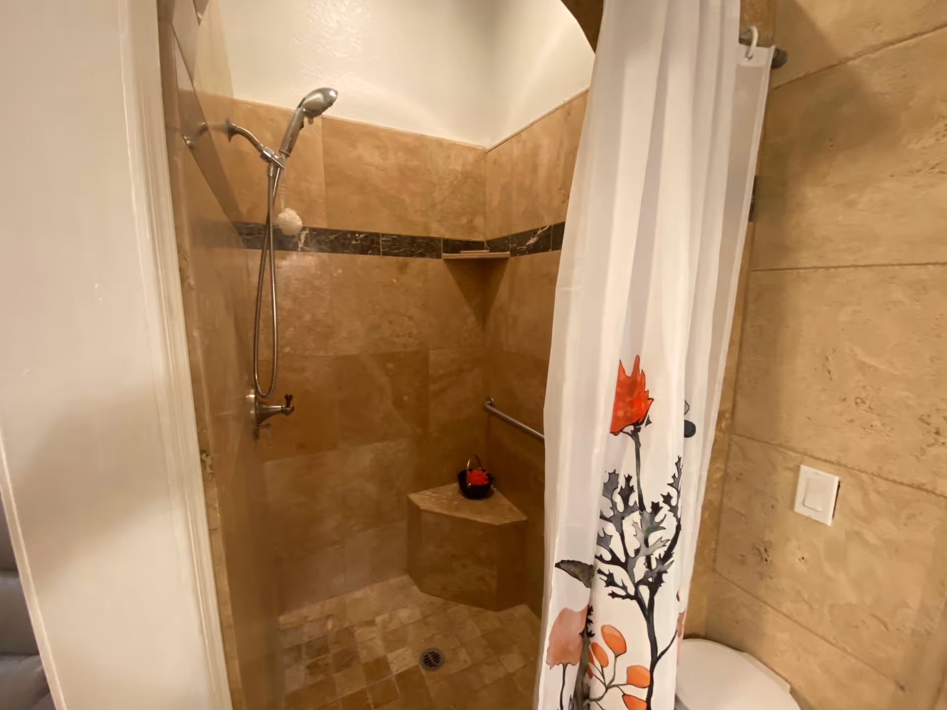 A tiled shower area with beige and brown tiles, a handheld showerhead mounted on the wall, a built-in corner bench, and a white shower curtain with a floral design partially drawn to the side.