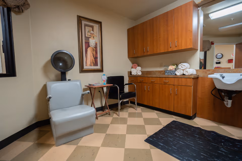 Interior room with a hair drying chair and hooded hair dryer, a black chair, a small table with magazines and a carton of milk, wooden cabinets with rolled towels on the countertop, a wall-mounted sink, a large mirror, and a framed painting of a woman wearing a hat.