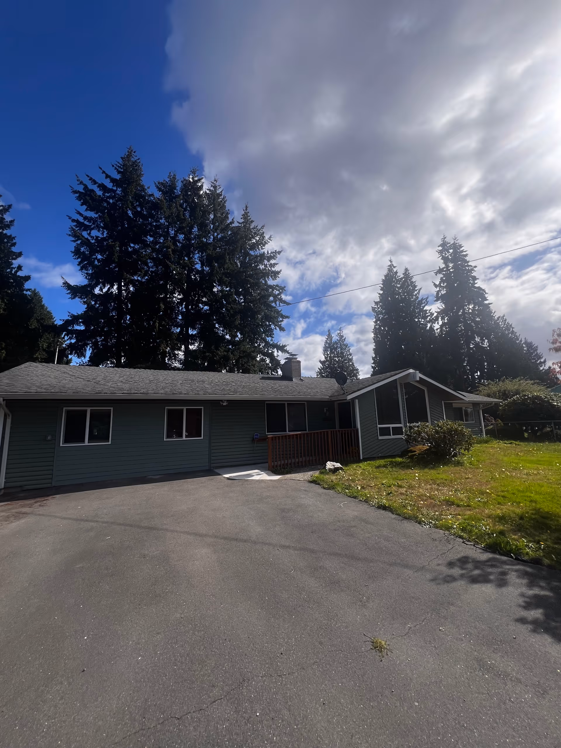Exterior view of a single-story house with driveway, front yard, and tall trees.