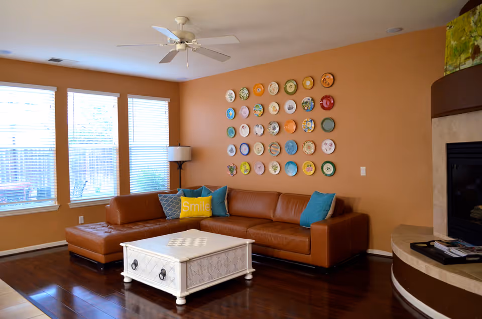 Bright living room with a brown leather sectional, decorative plates on the wall, a white coffee table, and large windows.