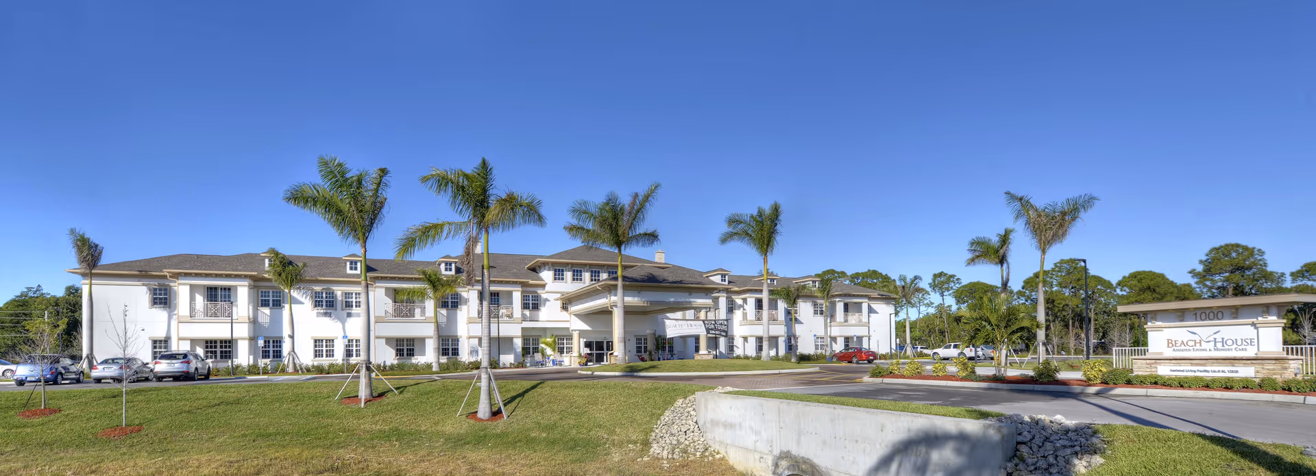Front exterior of a two-story white senior living building with palm trees, lawn, parked cars, and a driveway sign reading 'Beach House'.
