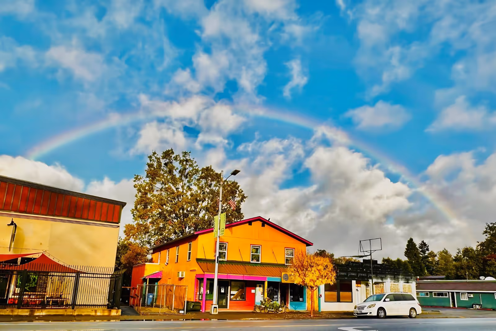 Vibrant rainbow arcs over colorful storefronts along a sunny street.