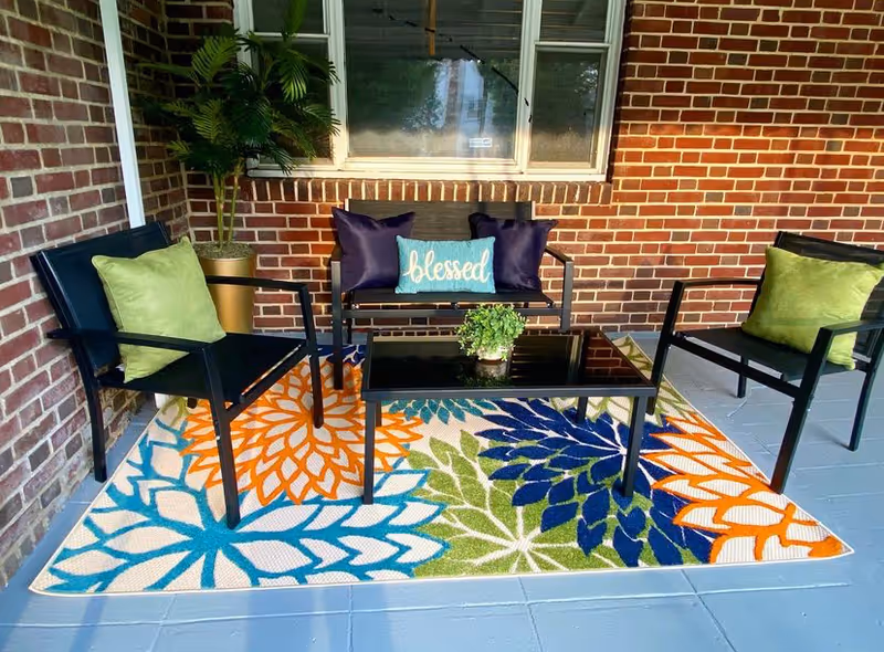 Outdoor brick porch with black metal seating, colorful rug, and potted plants.