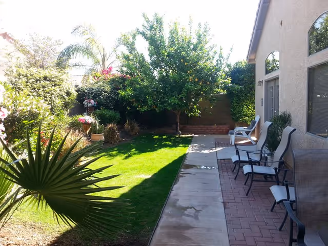 Sunlit outdoor patio with several chairs along a brick walkway beside a house and greenery.