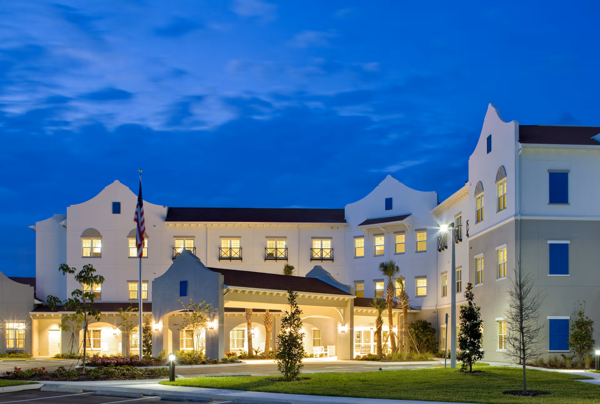 White, multi-story senior living building at dusk with illuminated entrance and flag.