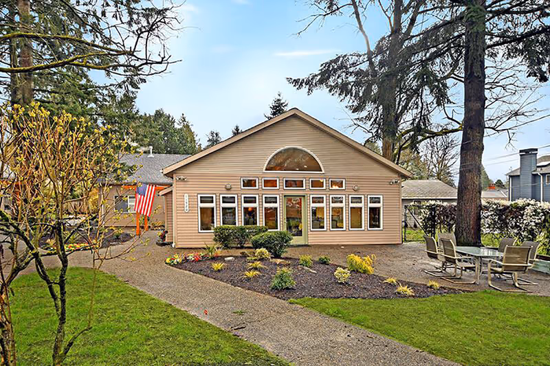 Front exterior of a single-story beige memory care building with large windows, landscaped grounds, an American flag, and outdoor seating.