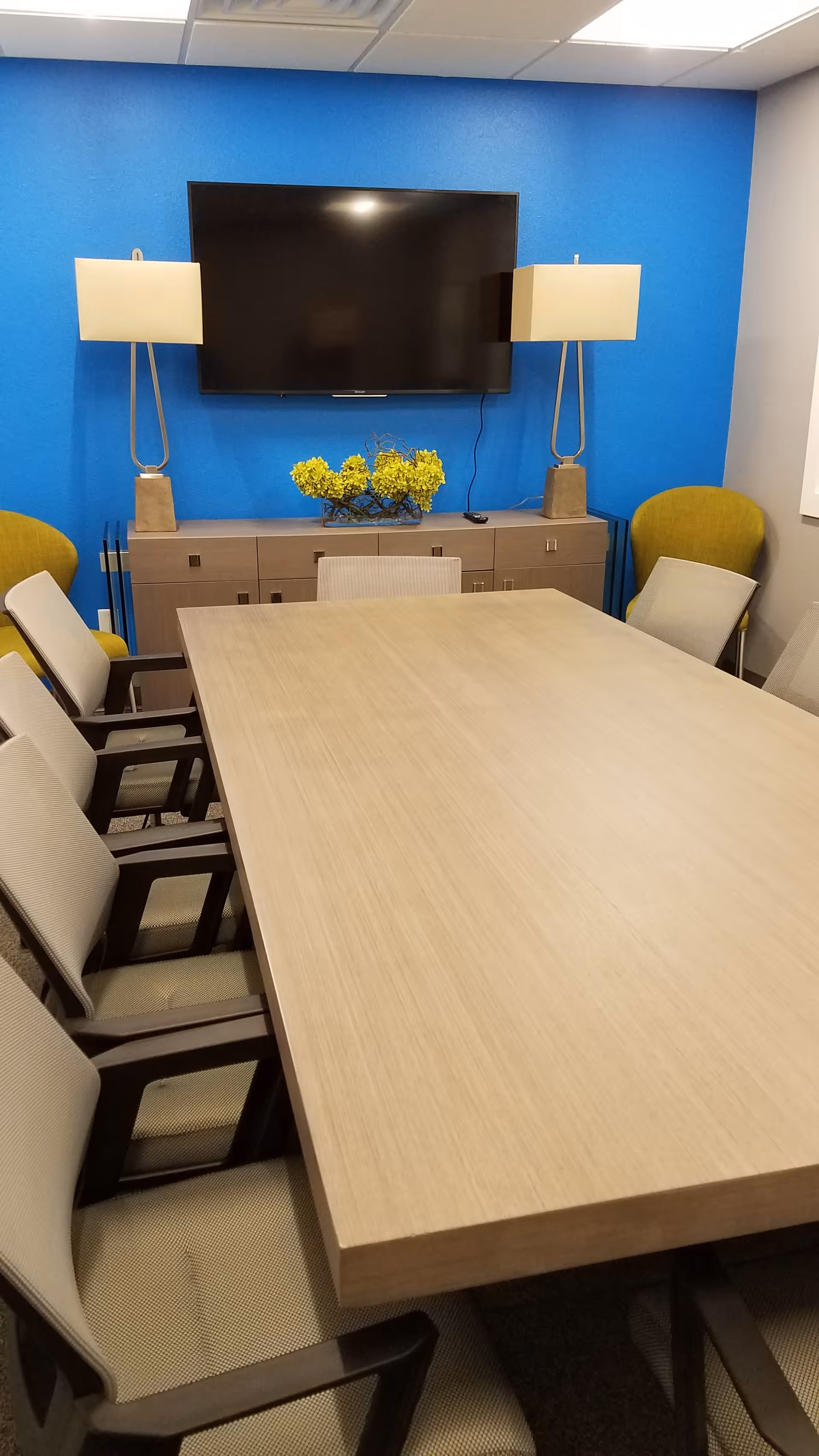 Conference room with a long table, beige chairs, blue accent wall, TV, and flowers.