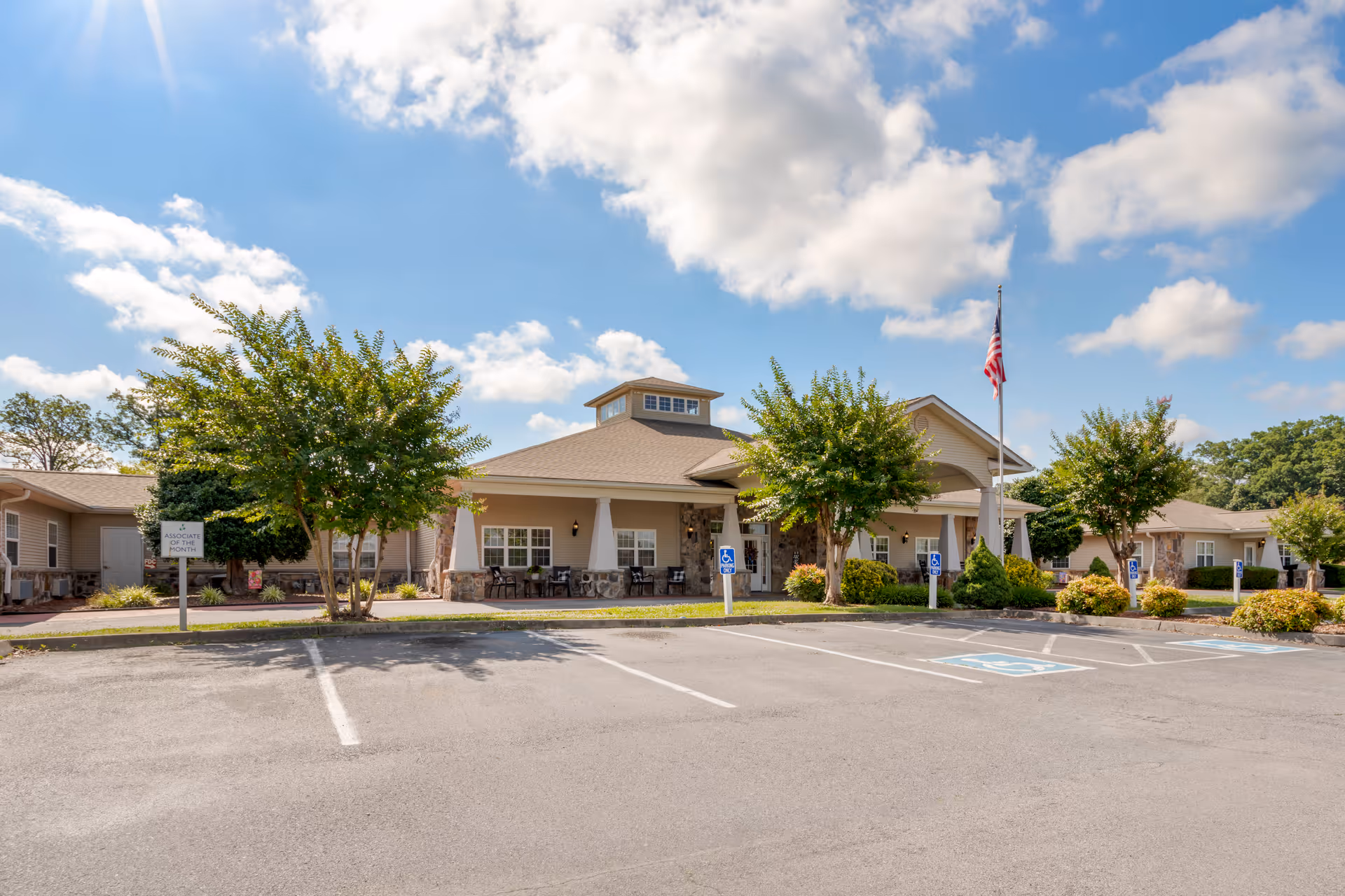Exterior view of a single-story senior living facility building with a covered entrance, several trees, and an American flag on a flagpole. There are multiple handicap parking spaces in front of the building under a partly cloudy sky.