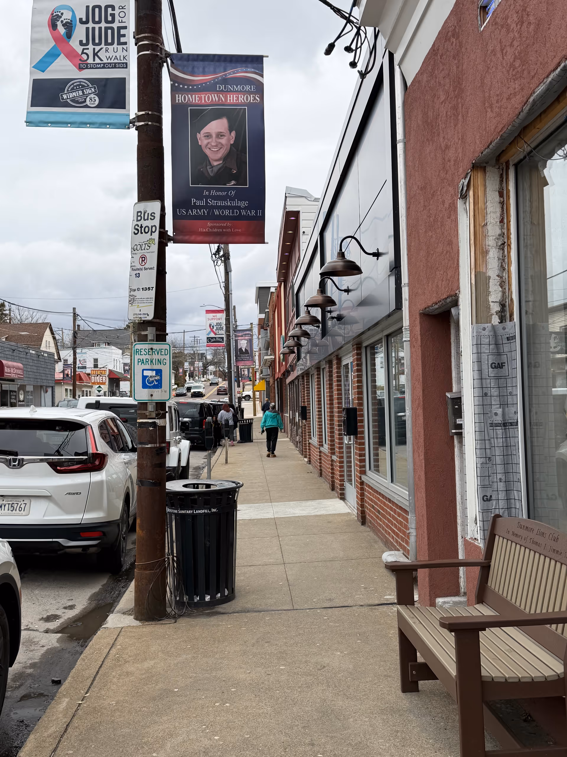 A city sidewalk with storefronts, banners, benches, and pedestrians along a sunny day.