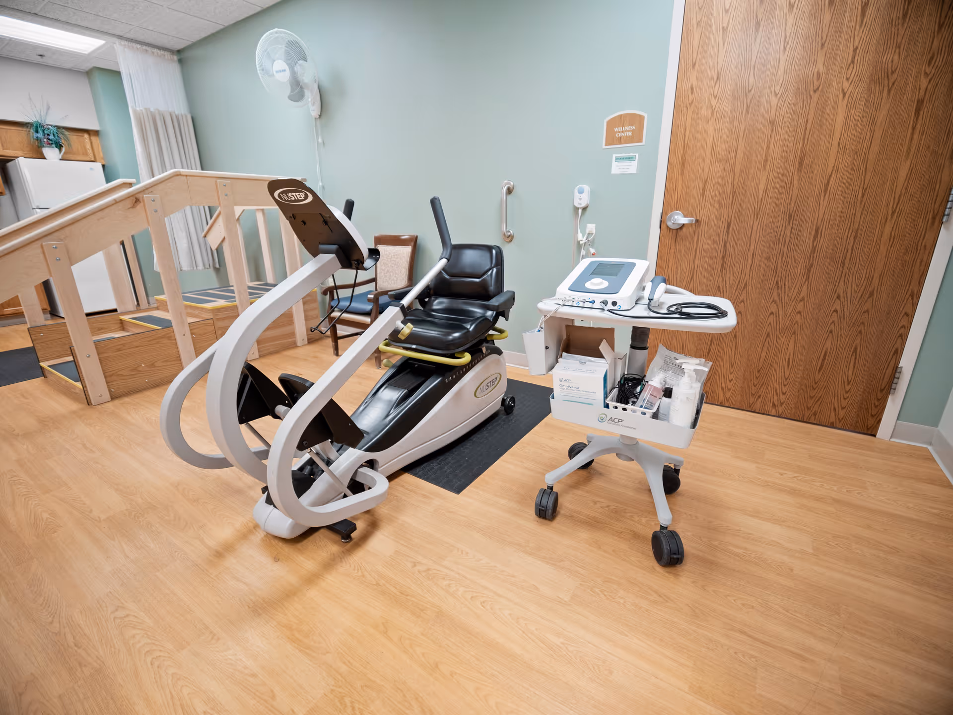 A small exercise and therapy room with a NuStep recumbent cross trainer machine, a medical equipment cart with various supplies, a wooden door labeled 'Wellness Center', a wooden ramp with handrails, and a wall-mounted fan. The room has light wood flooring and light green walls.