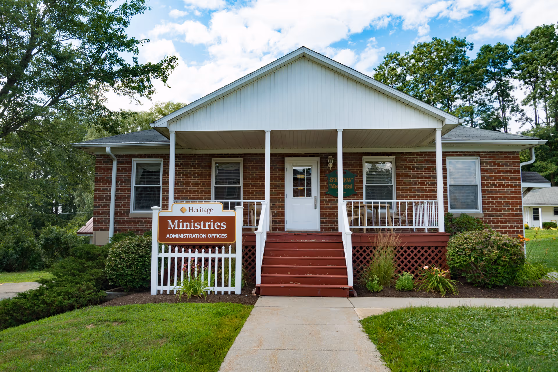 Brick adult home building with white front porch, steps, and a ministry sign.
