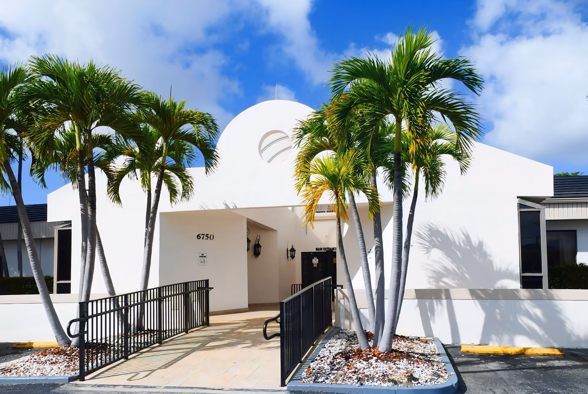 Modern white facility entrance with palm trees, ramps, and blue sky.